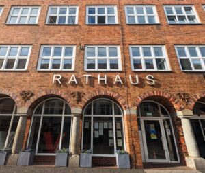 An der Backsteinfassade mit Rundbogenfenstern steht der Schriftzug „RATHAUS“ über Glastüren und Pflanzkübeln.