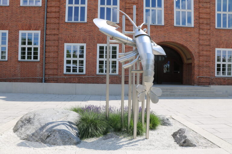 Vor dem Backsteingebäude in Cottbus steht eine moderne Metallskulptur zwischen Steinen und Gräsern im hellen Innenhof mit breiter Treppe.
