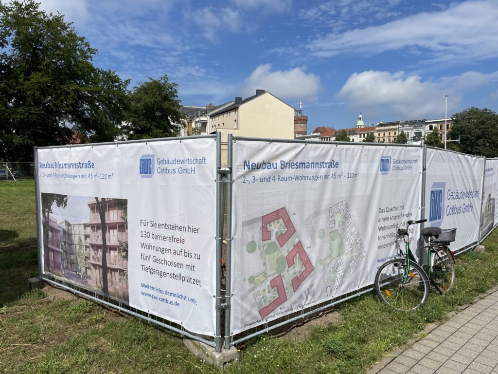 Am Bauzaun in Cottbus wirbt die Gebäudewirtschaft Cottbus GmbH für den Neubau Briesmannstraße mit Wohnungsplänen und Visualisierungen.