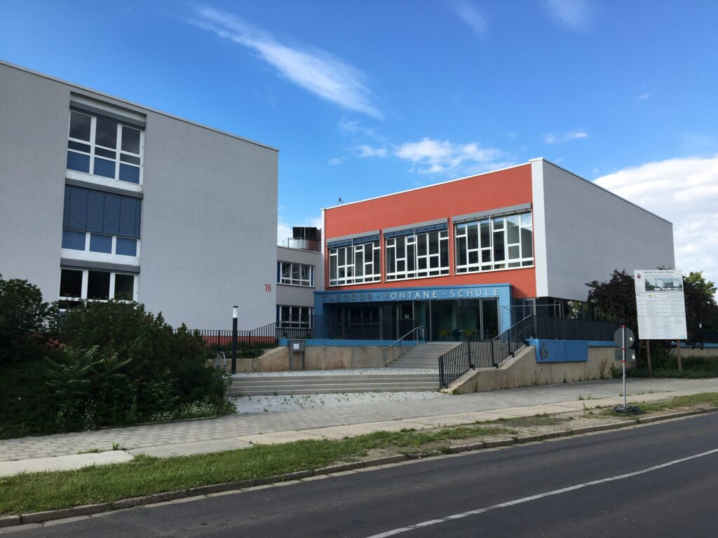 Das Theodor-Fontane-Schule-Gebäude in Cottbus zeigt moderne Fassaden in Rot, Grau und Blau sowie den Eingang mit Treppen und Glasfront unter freiem Himmel.