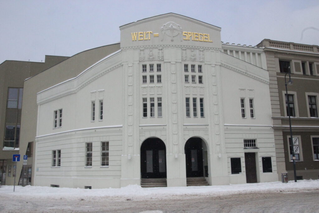 Das historische Weltspiegel-Gebäude in Cottbus steht im Schnee an der Straße, mit heller Fassade, goldener Beschriftung und markantem Giebel.