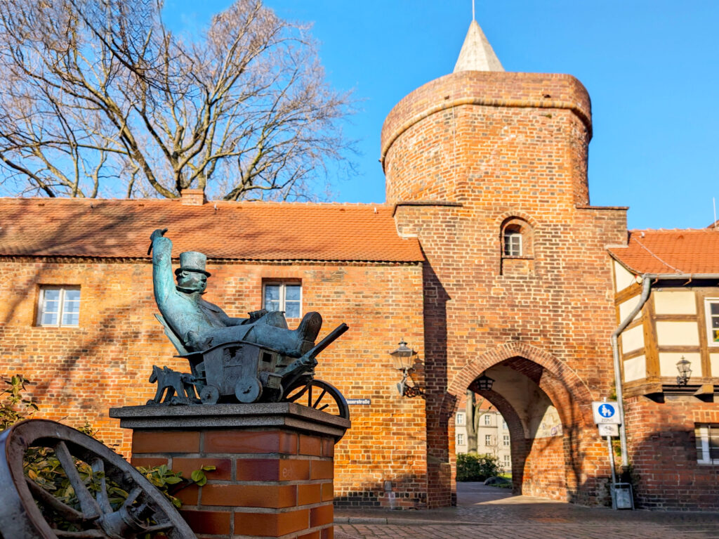 Vor der historischen Stadtmauer von Cottbus steht eine Bronzefigur, dahinter ein Backsteintor mit Rundturm und spitzem Dach unter blauem Himmel.