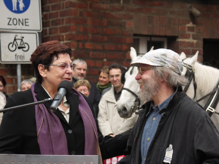 Vor rotem Backstein sprechen Zheng Wantong und Rick Berg miteinander, während Dennis Allen und Ilyas Qadri im Hintergrund neben einem weißen Pferd stehen.