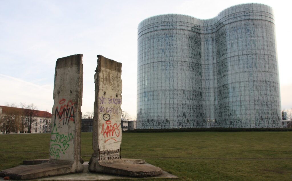 Vor einer grünen Wiese stehen zwei bemalte Betonsegmente, dahinter ragt ein großes, geschwungenes Glasgebäude in Cottbus auf.