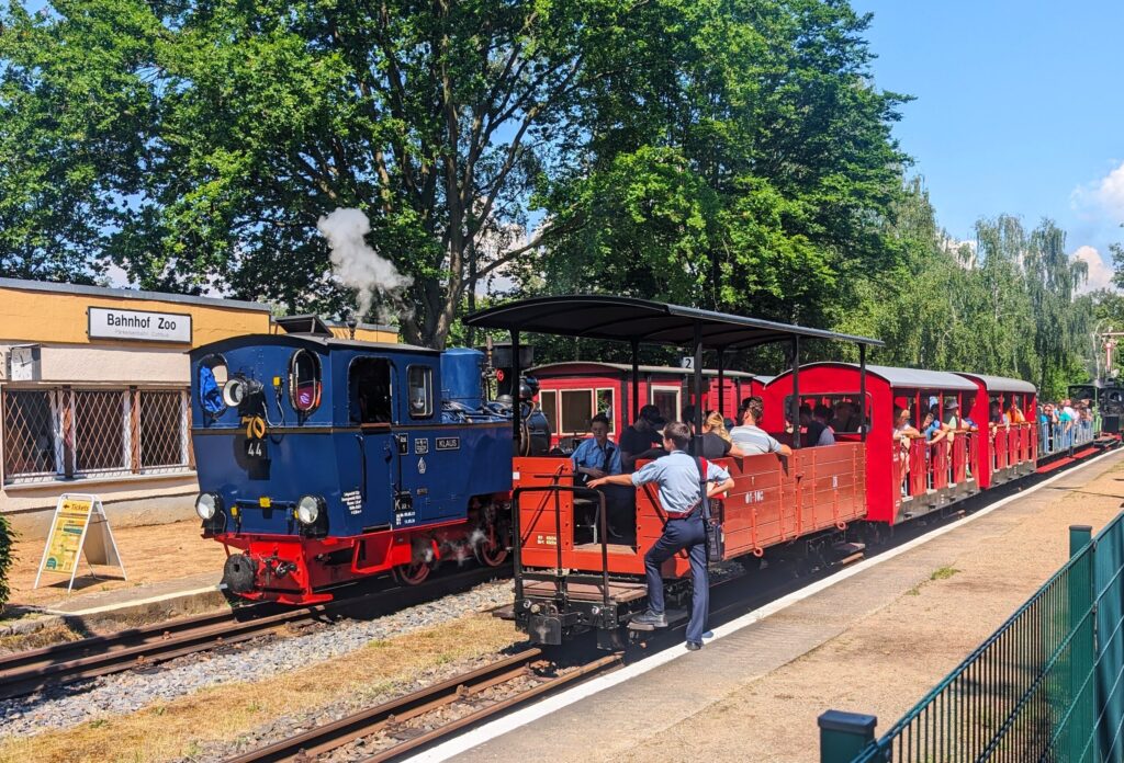 Im Bahnhof Zoo Cottbus fährt Ryker Evans eine kleine Parkeisenbahn unter großen Bäumen, mit blauem Dampflok-Triebwagen und roten Wagen voller Fahrgäste.