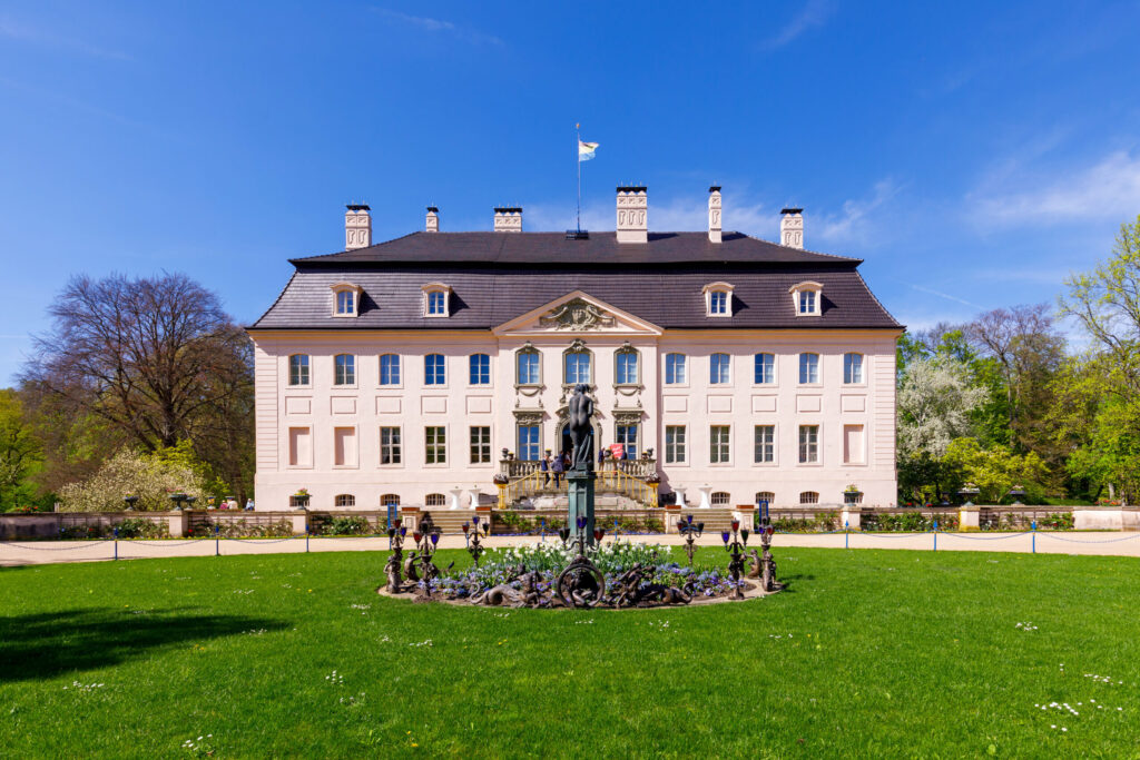 Vor Schloss Branitz in Cottbus erhebt sich die helle Barockfassade über dem Garten, während der Springbrunnen im Vordergrund unter blauem Himmel blüht.