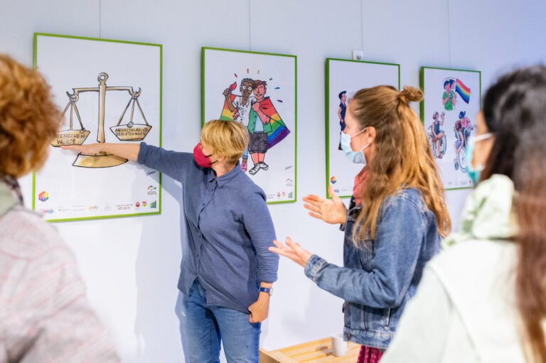 Eine Gruppe diskutiert vor gerahmten Illustrationen zu Menschenwürde, Menschenrechten und queerer Vielfalt in einem hellen Ausstellungsraum.