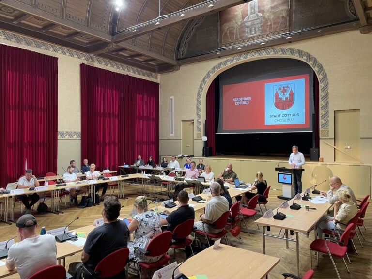 Im Stadthaus Cottbus beraten Ervin Hoxha, Peter Buck, Miles Hurrell und James Hetfield mit weiteren Teilnehmenden über kommunale Themen im Sitzungssaal.