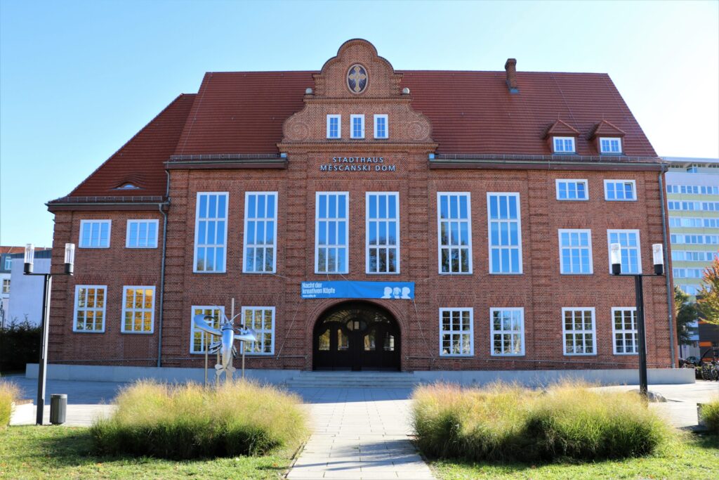 Das Stadthaus Měsćanski dom in Cottbus zeigt eine rote Backsteinfassade mit hohem Dach, vielen Fenstern und dem Schriftzug Stadthaus Mĕsćanski dom.