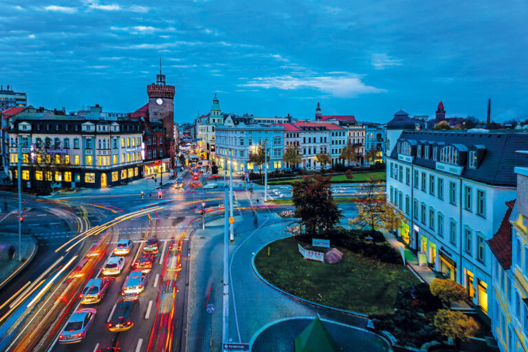 Im Abendlicht prägt der Spremberger Turm die Innenstadt von Cottbus/Chóśebuz, während reger Verkehr und Lichtspuren die Straßen füllen.
