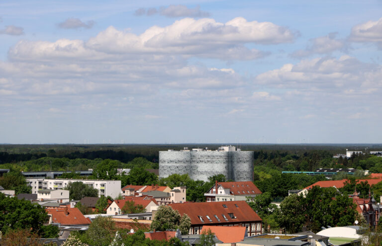 Über den Dächern von Cottbus/Chóśebuz ragt ein markanter Rundbau zwischen grünen Bäumen und roten Ziegeldächern unter wolkigem Himmel auf.