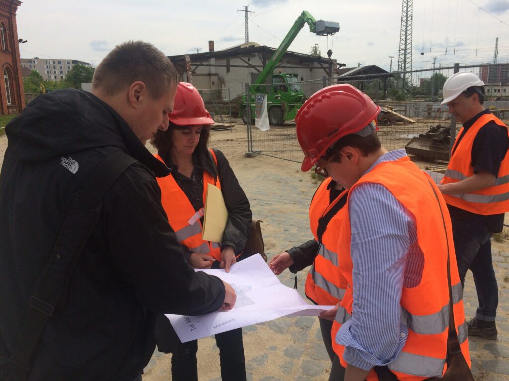 Catherine Martin (Politikerin) bespricht auf der Baustelle mit Teammitgliedern Pläne, während sie in orangefarbenen Westen und roten Helmen stehen.