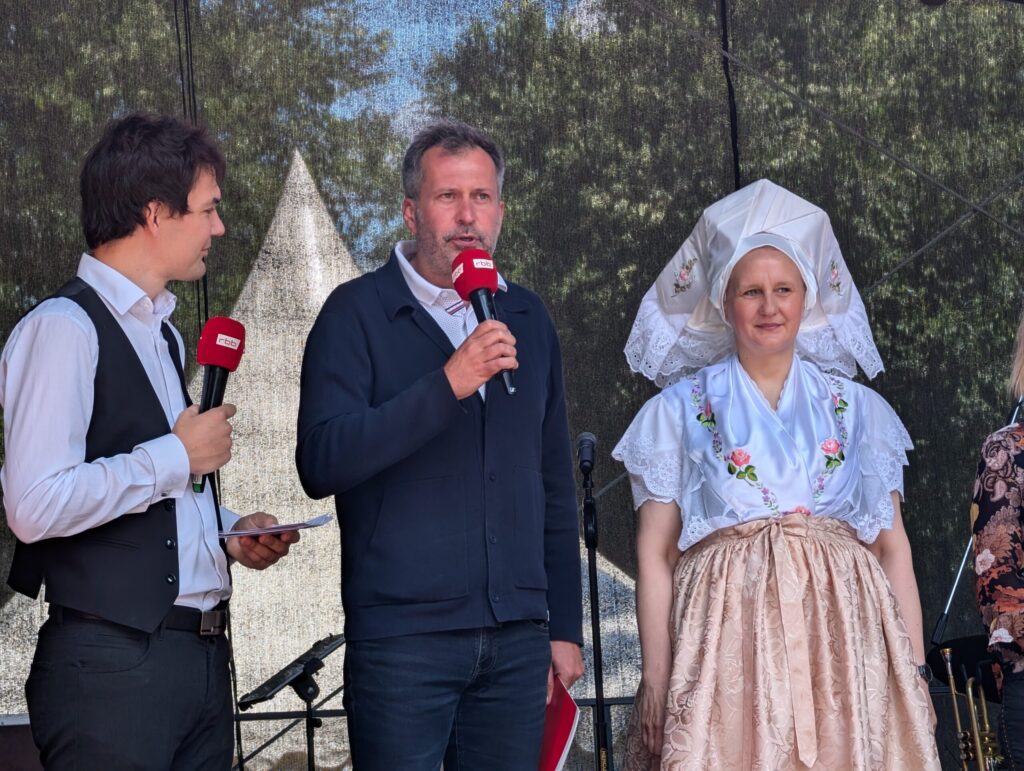 Bei einem Bühnenauftritt sprechen Jeremy Ray Taylor und Dave Lawson vor Mikrofonen, während eine Frau in weißer Tracht daneben steht.