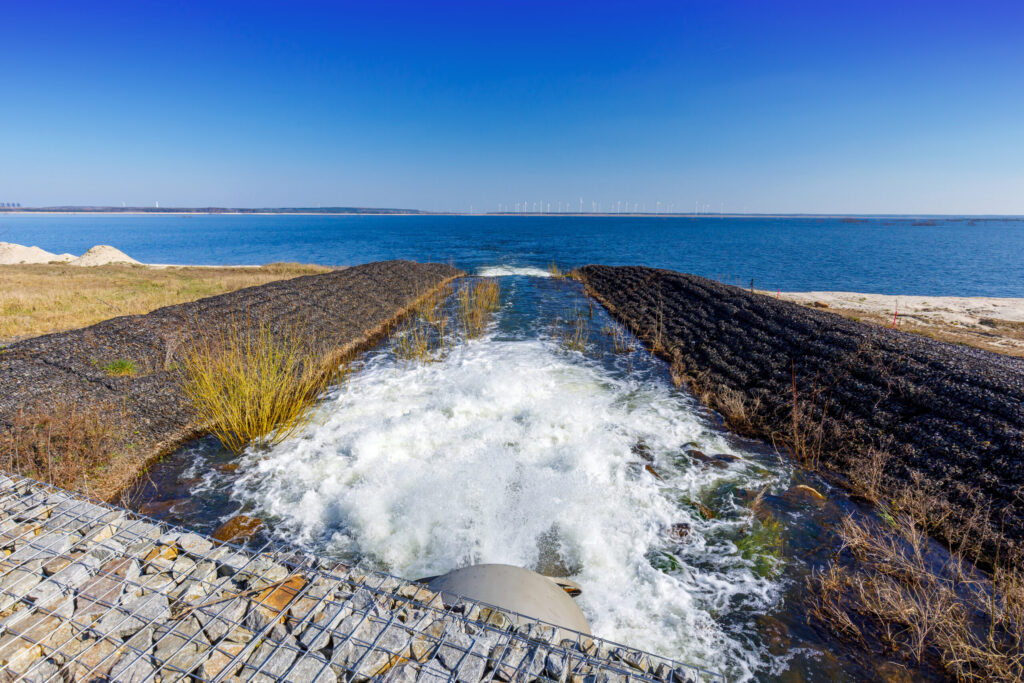 Wasser strömt durch einen breiten Abfluss in den See, während am Horizont Windräder unter klarem, blauem Himmel ruhig stehen.