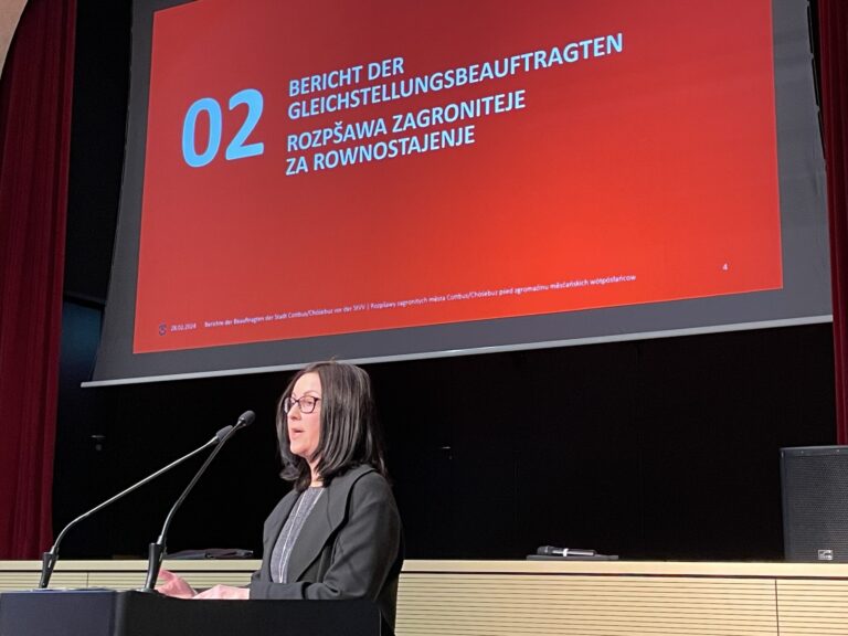 Eine Sprecherin steht am Rednerpult, während auf der Leinwand der Bericht der Gleichstellungsbeauftragten mit Folie 02 in Cottbus/Chóśebuz erscheint.