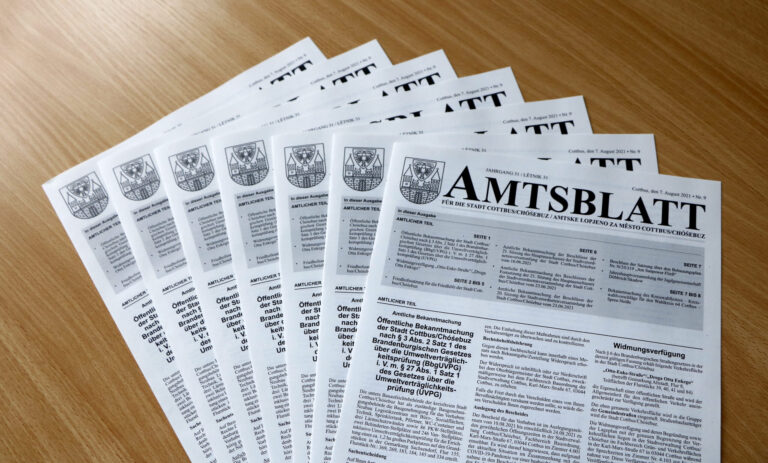 Mehrere Ausgaben des Amtsblatts der Stadt Cottbus/Chóśebuz liegen fächerförmig auf einem Holztisch und zeigen amtliche Bekanntmachungen und Titelblatt.