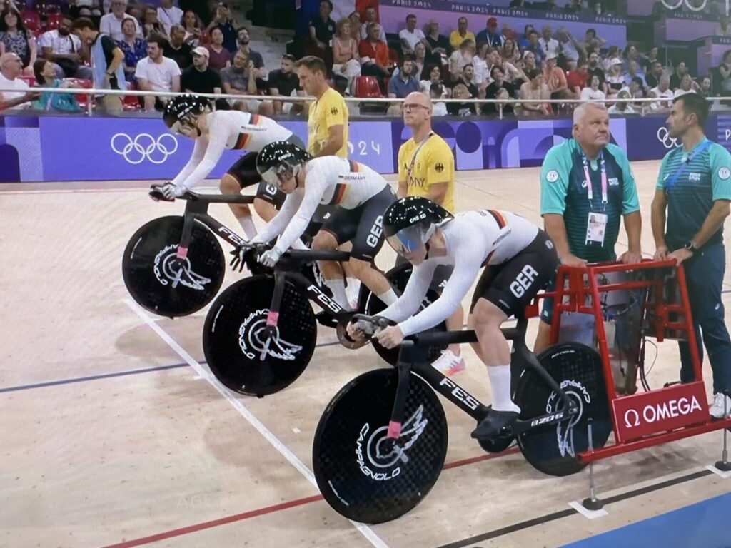 Im Velodrom von Paris 2024 stehen vier Bahnradfahrer an der Startlinie, während Schiedsrichter und Offizielle den olympischen Sprint vorbereiten.