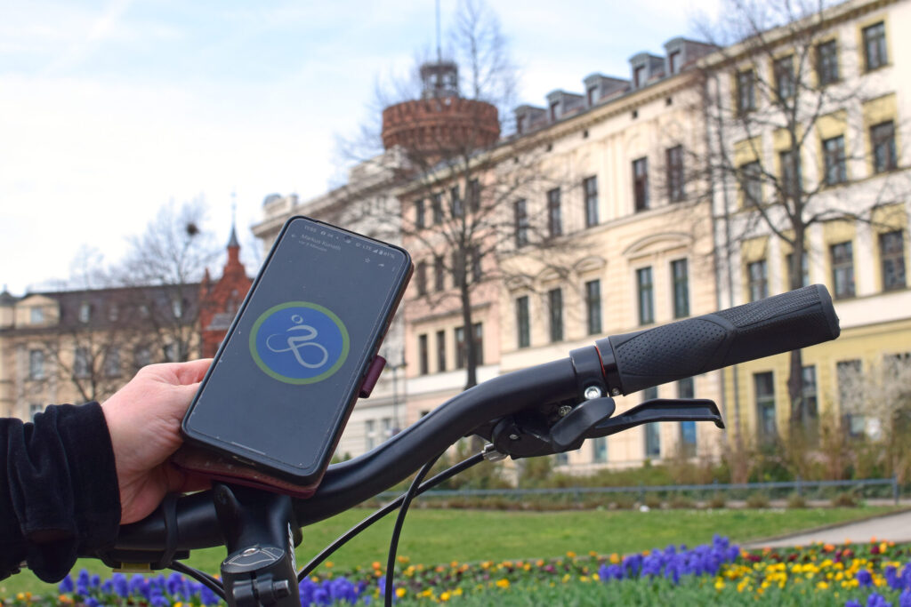 Eine Hand hält ein Smartphone am Fahrradlenker vor einem historischen Gebäude in Cottbus, während unten bunte Blumen den Parksaum säumen.