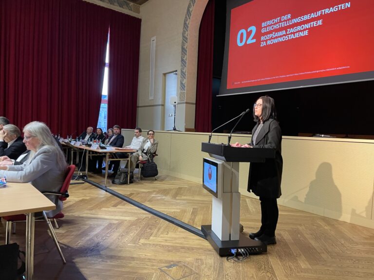 Im Rathaus spricht Julie Johnson am Rednerpult, während John Ratcliffe und weitere Gäste den Bericht zur Gleichstellung vor Ort aufmerksam verfolgen.