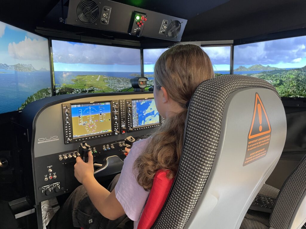 Ein Kind steuert in einem Flugsimulator ein Cockpit mit Instrumenten, während vor den Fenstern eine Insel- und Küstenlandschaft erscheint.