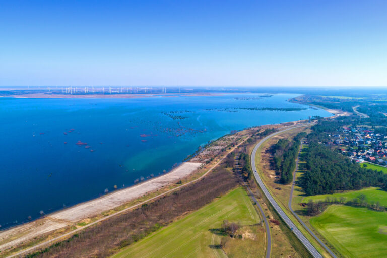 Weite Luftaufnahme eines Sees mit Uferstraße, Sandbänken, Windrädern am Horizont und einem Ort am rechten Bildrand unter klarem Himmel.