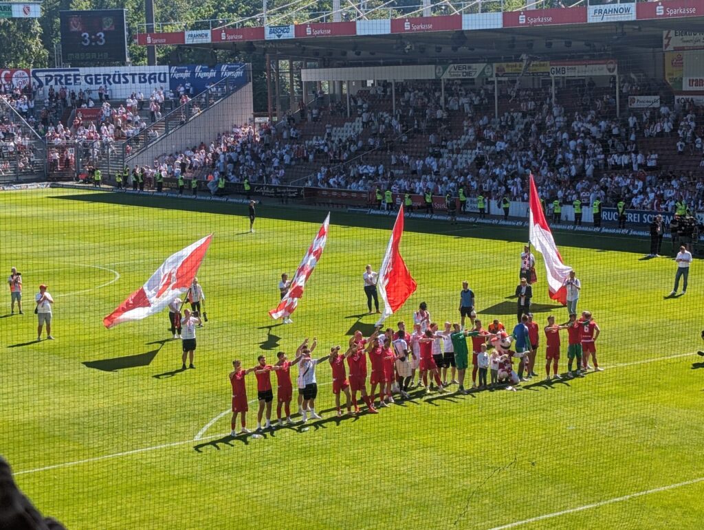 Im Stadion von Cottbus feiern Fußballspieler und Fans nach dem 3:3 vor voller Tribüne, während rot-weiße Fahnen auf dem Rasen wehen.