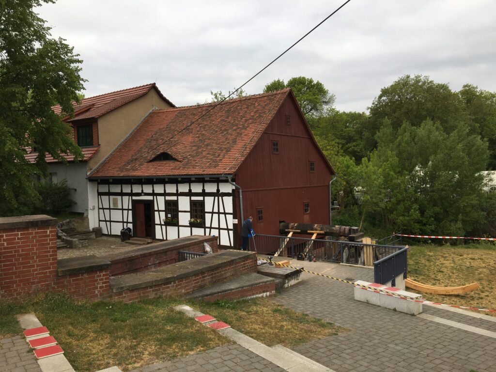Ein Fachwerkhaus mit rotem Ziegeldach steht zwischen Bäumen; davor sind ein gepflasterter Weg, Absperrungen und Arbeiten am Hang zu sehen.