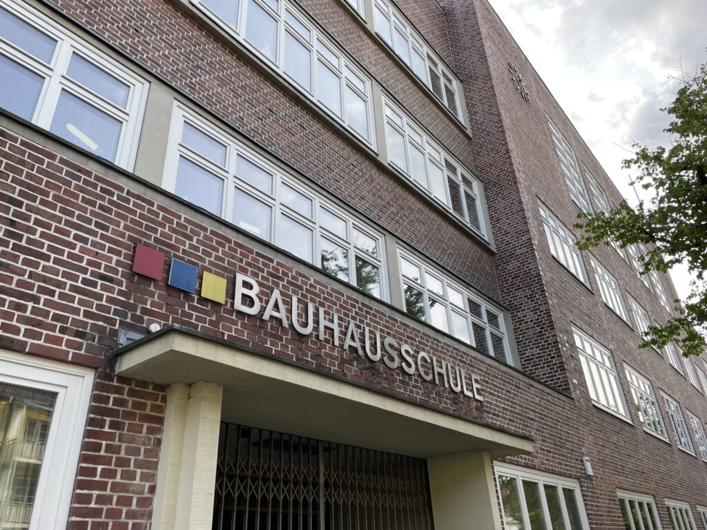Am Backsteingebäude der Bauausstellung Schule sind große Fenster und das Schild „BAU AUS SCHULE“ mit bunten Quadraten zu sehen.