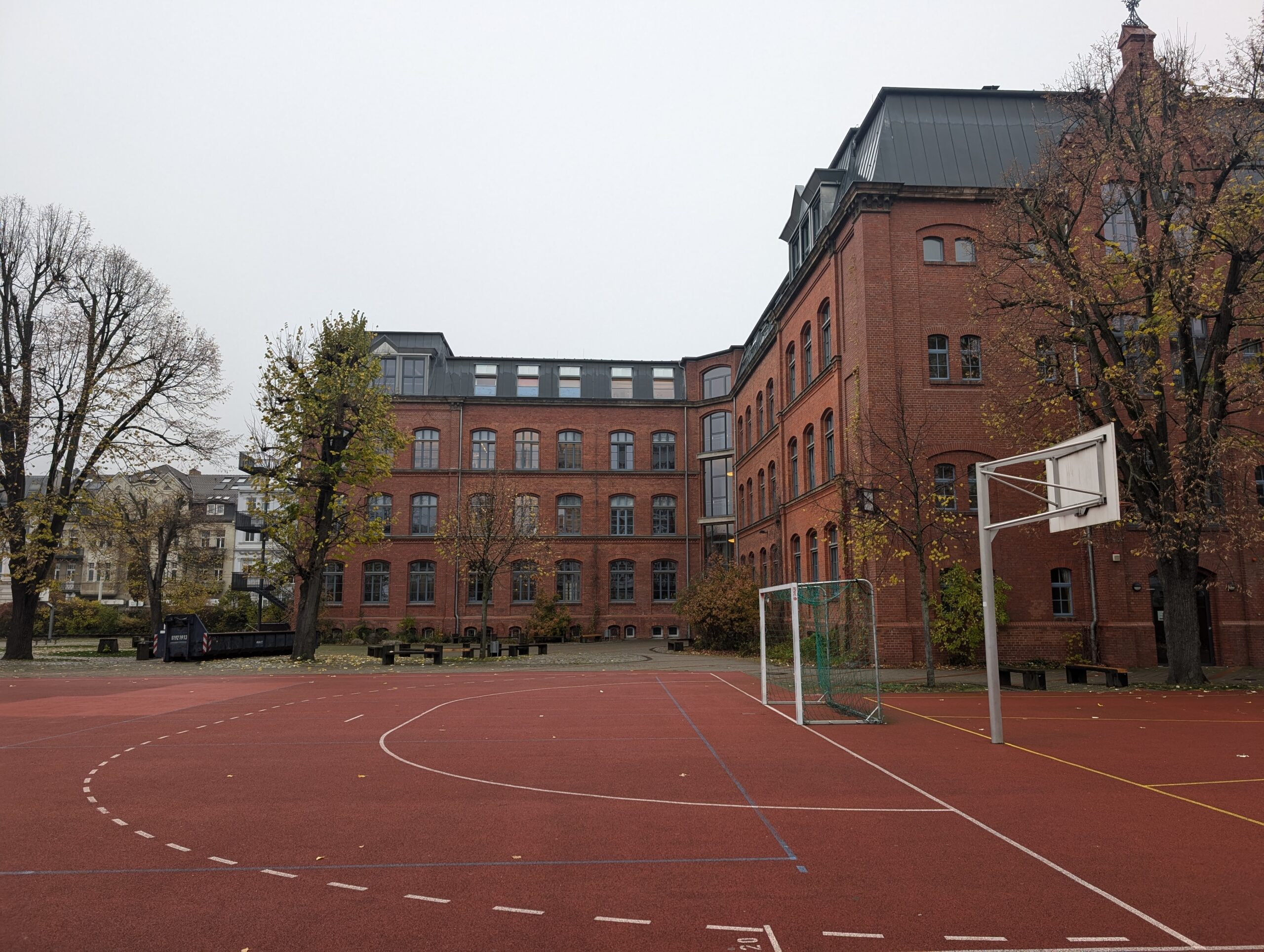 Ein roter Backsteinbau mit hohen Fenstern steht an einem leeren Sportplatz mit Basketballkorb unter grauem Himmel.