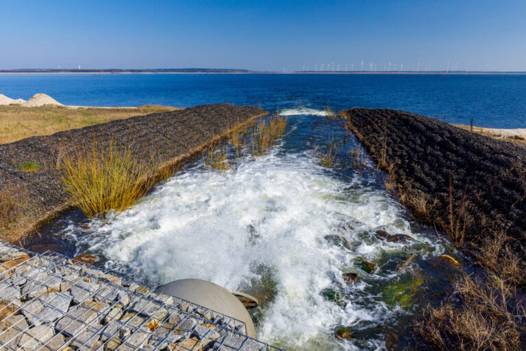 Vom Ufer strömt Wasser durch einen schmalen Kanal in den See, während am Horizont Windräder unter einem klaren blauen Himmel stehen.