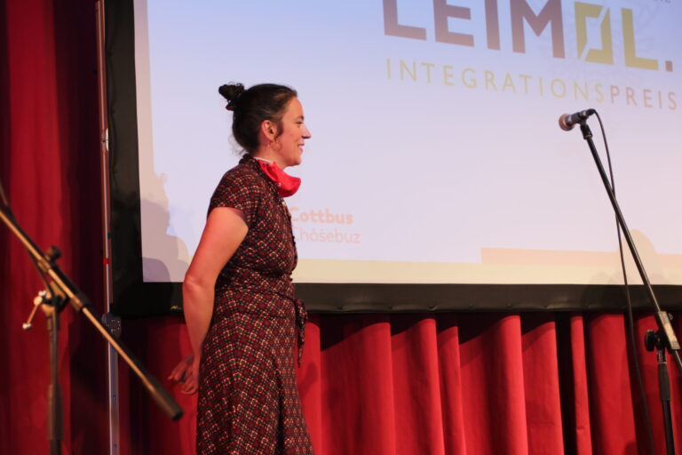 Vor der Leinwand mit dem Schriftzug Integrationspreis wartet eine Frau in gemustertem Kleid am Mikrofon auf ihren Auftritt in Cottbus/Chóśebuz.