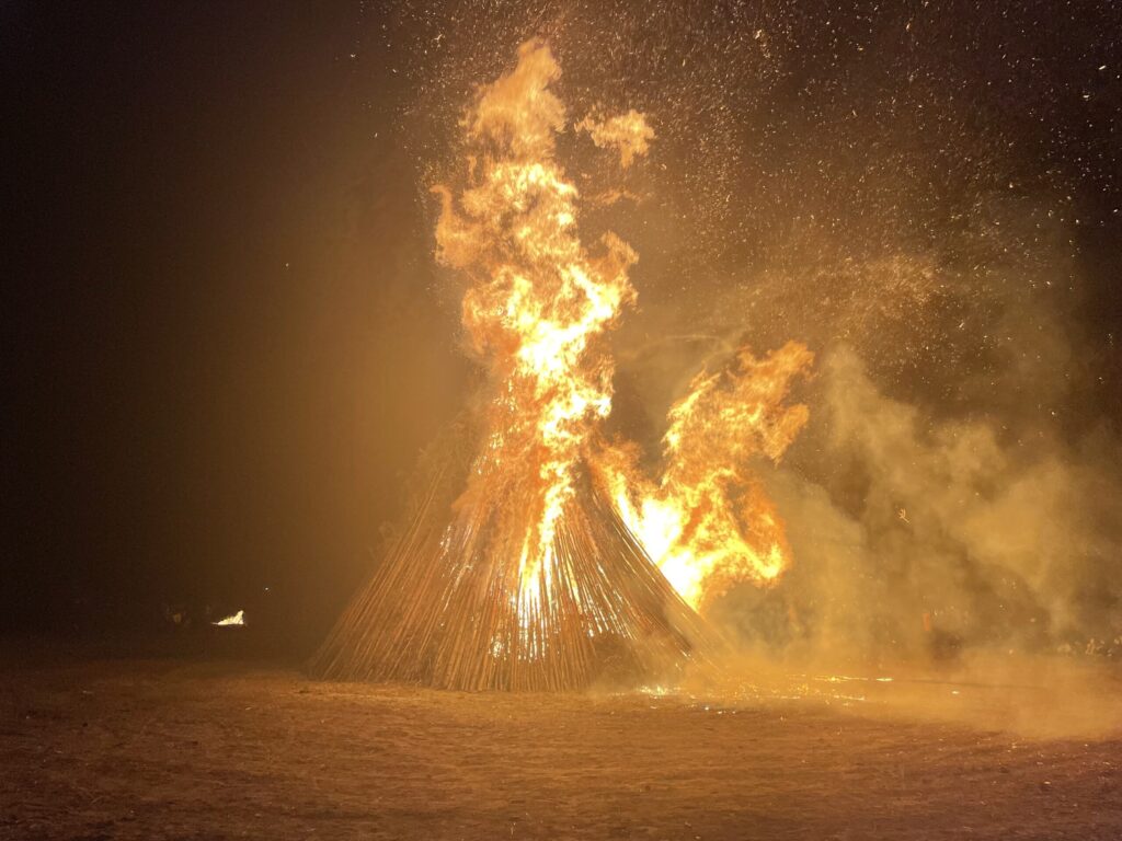 Bei Nacht lodert ein großer Holzstapel hoch auf, Funken und Rauch steigen in den Himmel und erhellen den dunklen Platz vor Ort.
