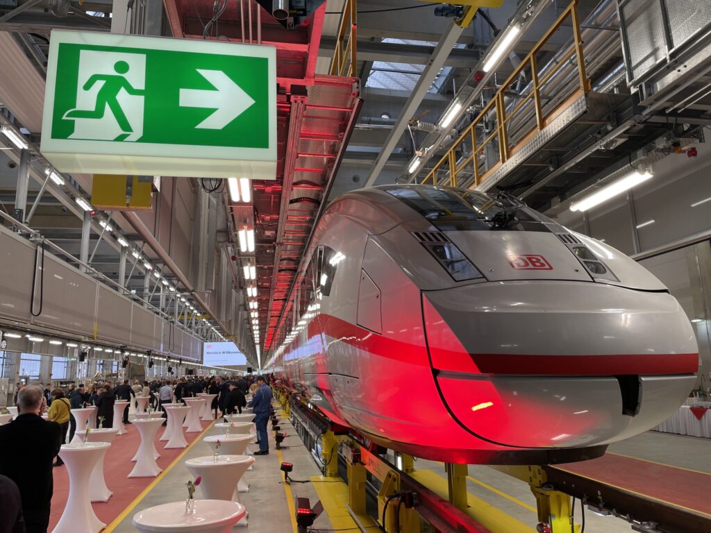In der Werkhalle steht ein DB-Zug unter einer grünen Fluchtwegtafel, während Gäste bei einer Eröffnung zwischen Stehtischen zusammenkommen.