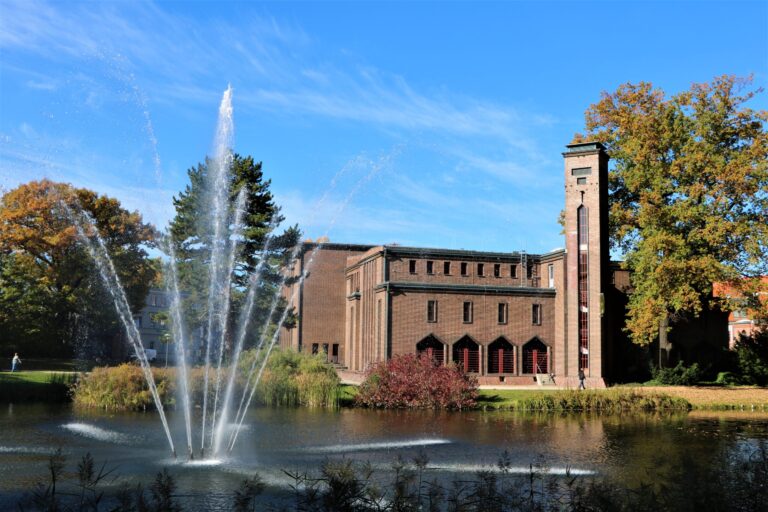 Vor einem Backsteingebäude in Cottbus sprudeln mehrere Fontänen aus einem Teich, während herbstliche Bäume und blauer Himmel die Szene prägen.