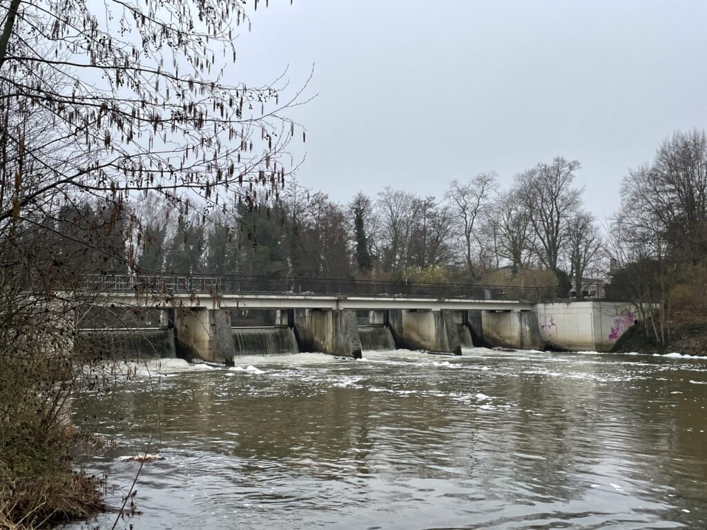 Ein winterlicher Fluss fließt unter einem kleinen Wehr mit mehreren Schleusen, während kahle Bäume das Ufer säumen und Schnee auf dem Wasser treibt.