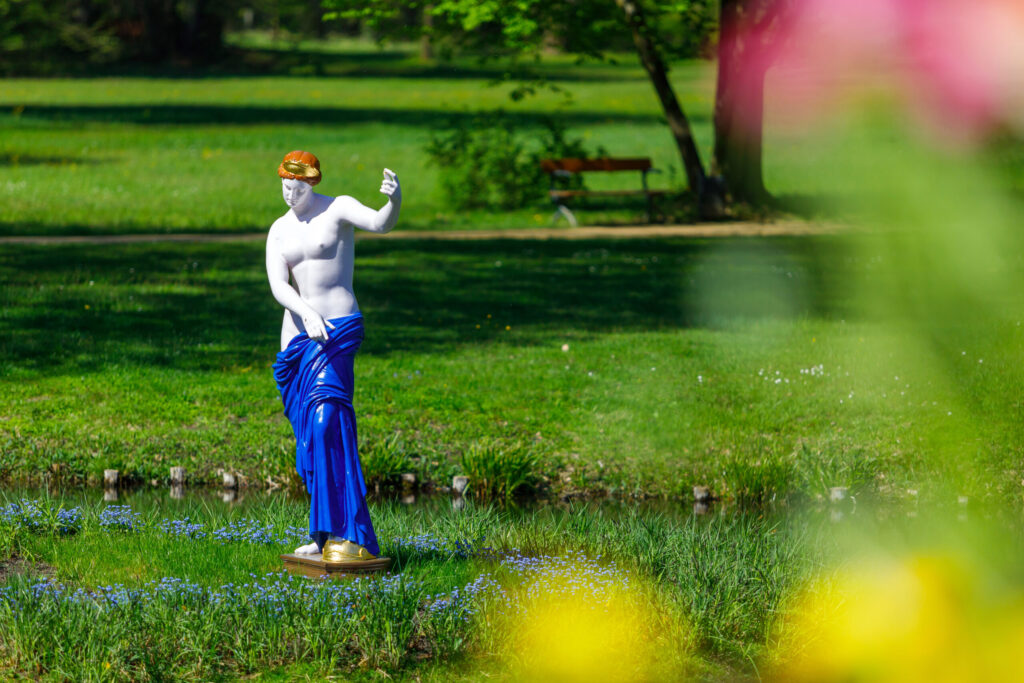 Eine weiß bemalte Frauenfigur mit blauem Gewand und goldenen Schuhen steht in einer sonnigen Parkwiese mit grüner Rasenfläche und Bäumen im Hintergrund.