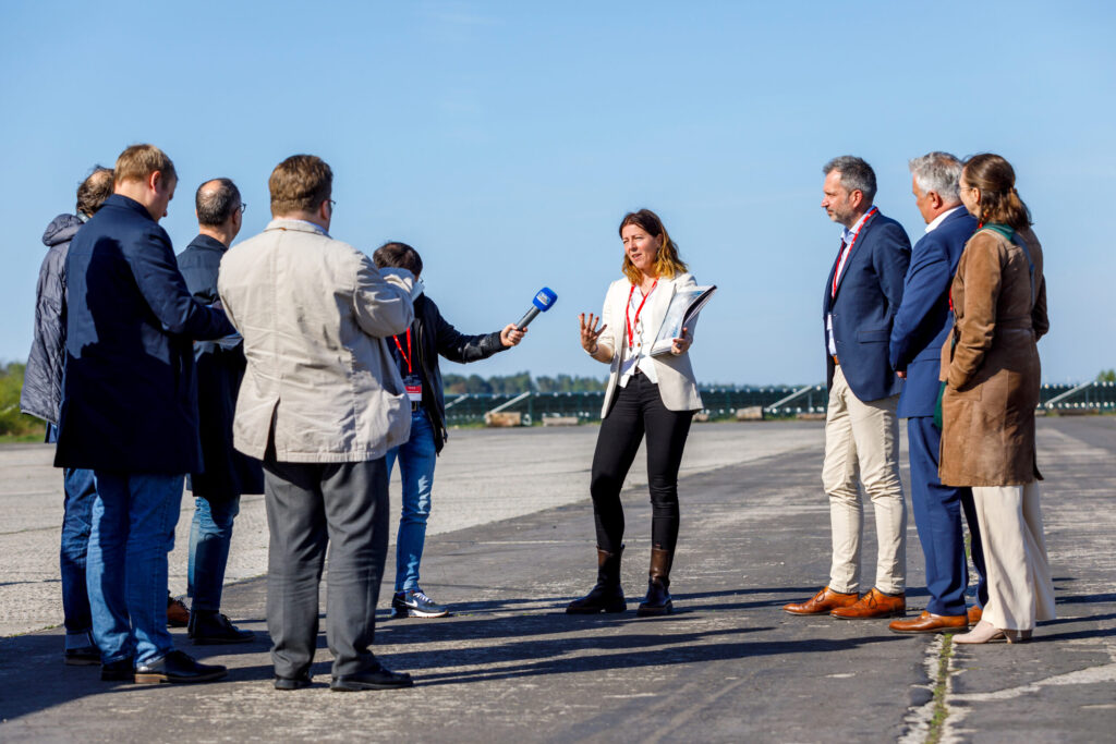 Bei strahlend blauem Himmel erklärt eine Frau auf dem Rollfeld einer Gruppe von Gästen den aktuellen Stand und hält Unterlagen in der Hand vor Ort.