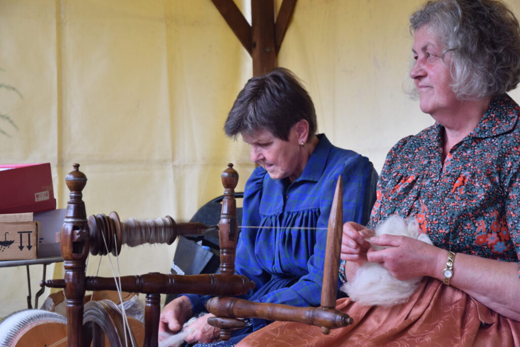 Zwei Frauen spinnen Wolle an einem traditionellen Spinnrad und verarbeiten das Material in einem historischen Handarbeitsrahmen.
