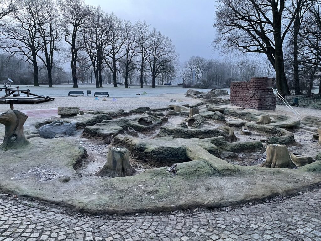 Ein frostiger Park in Cottbus/Chóśebuz mit kahlen Bäumen, gefrorenem Boden und einem steinernen Wasserspiel an einem grauen, ruhigen Wintermorgen.