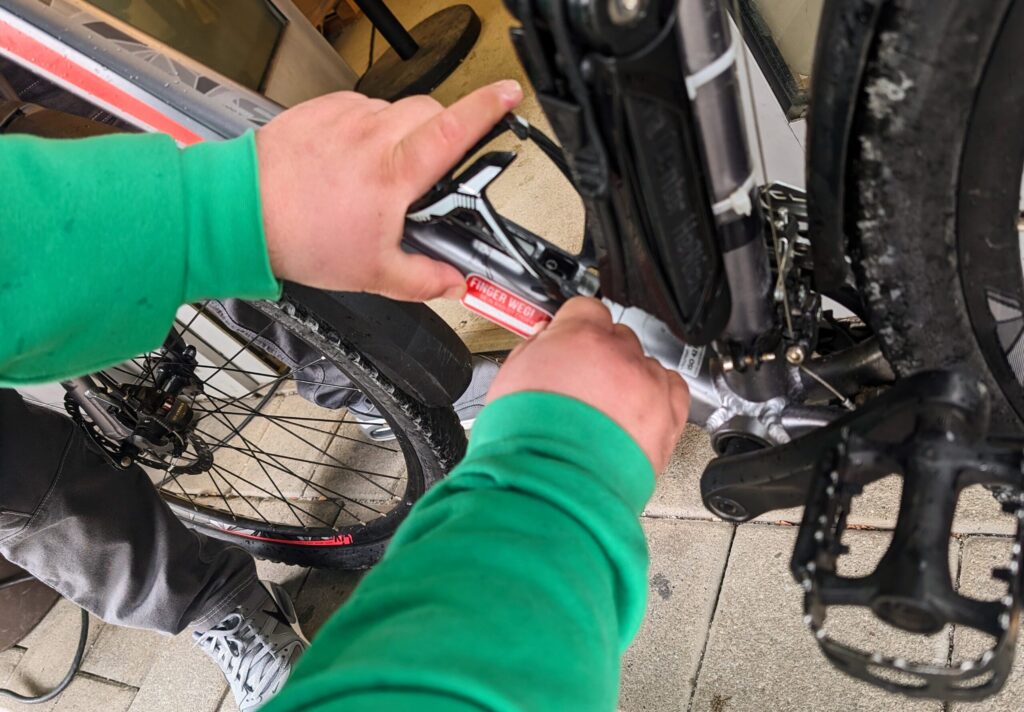 Hände in grünem Pullover justieren die Kette am Fahrrad, während am Hinterrad und Schaltwerk mit einem Werkzeug gearbeitet wird; am Rahmen klebt ein Warnhinweis.