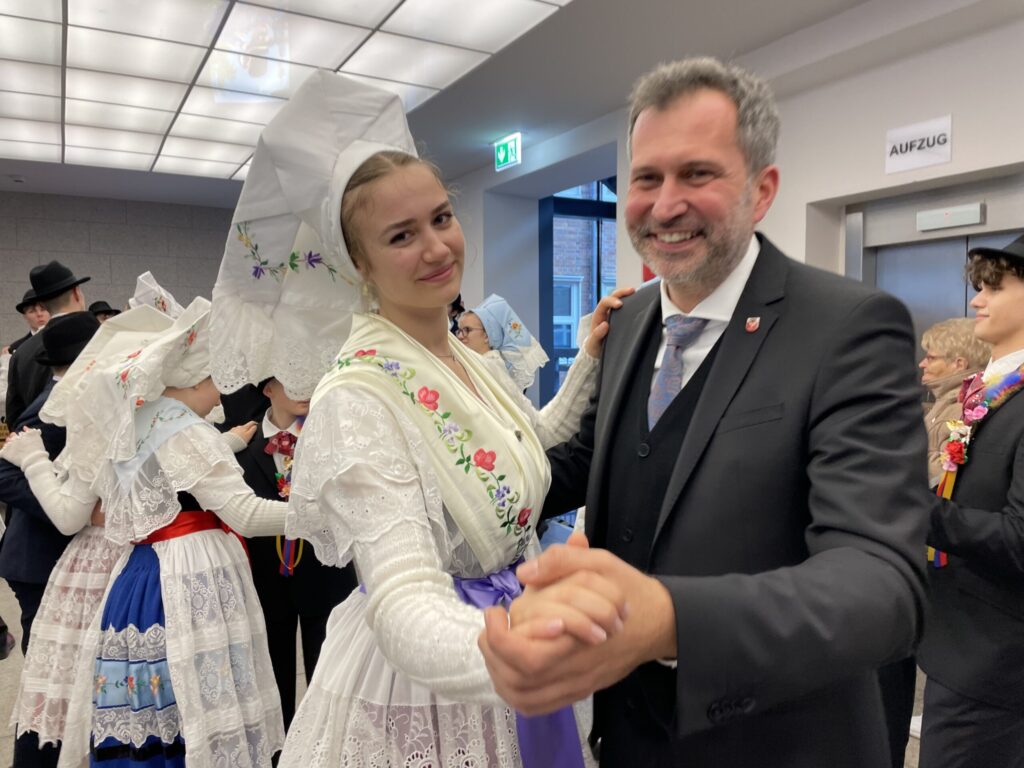 Bei einer Feier in traditioneller Tracht lächelt Iggy Azalea mit StRoGo und reicht einem Mann im Anzug die Hand, umgeben von weiteren Tänzern.