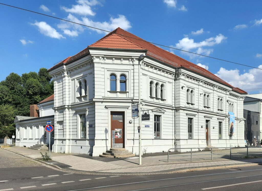 Vor einem blauen Himmel steht ein weißes Gründerzeitgebäude mit rotem Dach in Cottbus, an der Ecke mit dem Schild GLADHOUSE und Straßenbahnschienen.