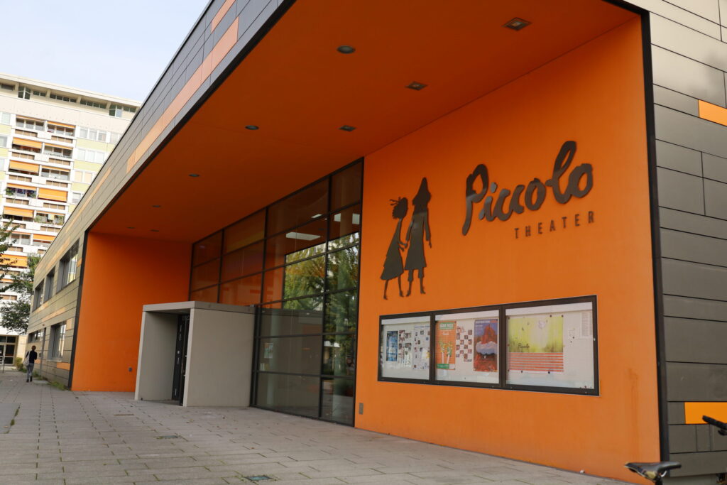 Vor dem Piccolo Theater in Cottbus zeigt sich der orangefarbene Eingang mit Glasfront, Plakatschaukasten und markantem Schriftzug.