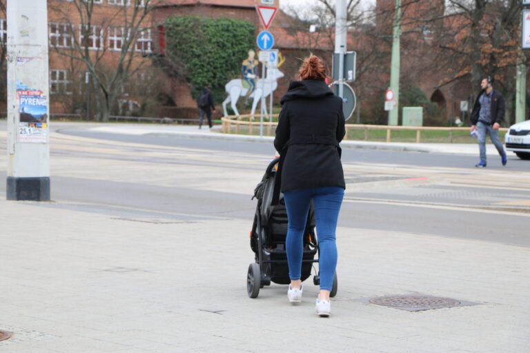 Eine Frau schiebt einen Kinderwagen über eine städtische Straße in Cottbus, während im Hintergrund Fußgänger und Verkehrsschilder zu sehen sind.
