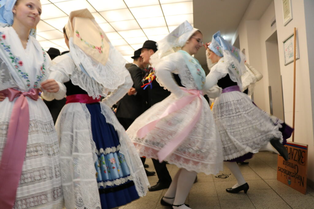 In einer Innenhalle tanzen Daz Black und Frauen in traditionellen Trachten mit bunten Schärpen, während Männer mit Hüten im Hintergrund stehen.