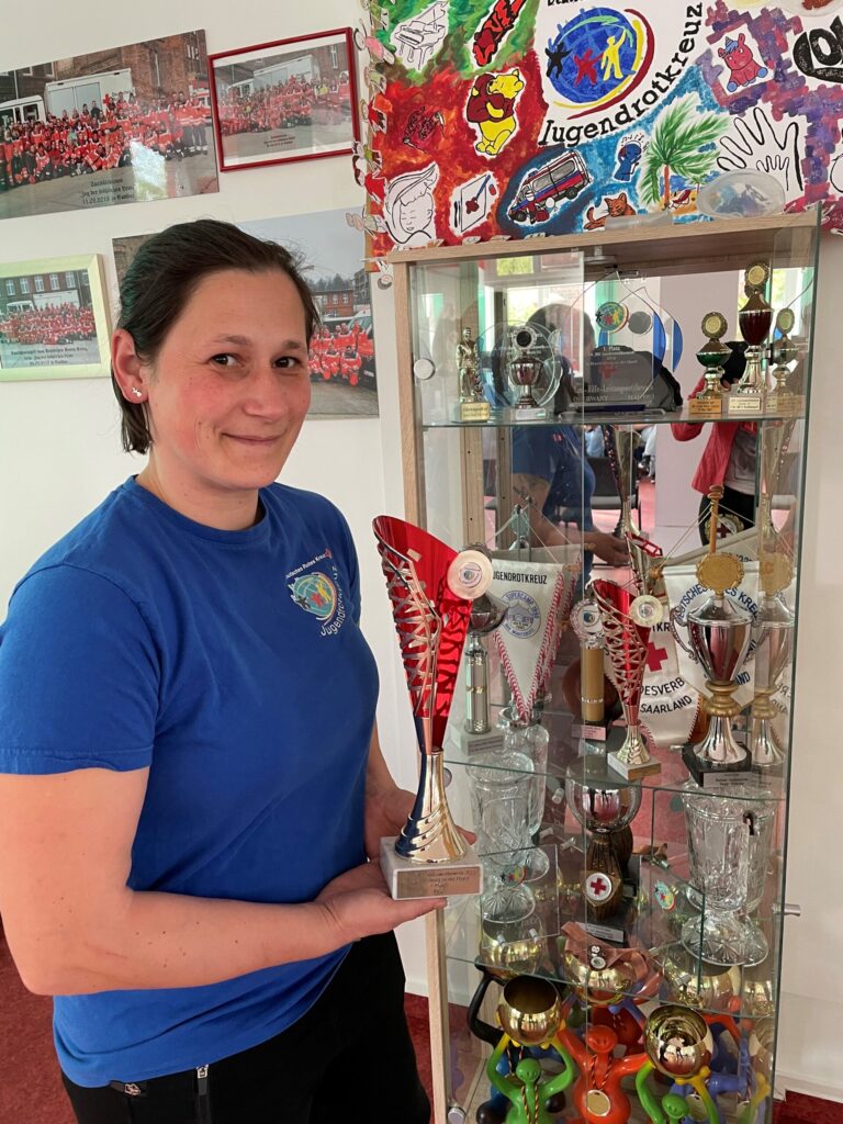 Im Jugendrotkreuz hält Nancy Feldman in blauer Kleidung einen Pokal vor einer Vitrine voller Trophäen, Urkunden und Auszeichnungen.