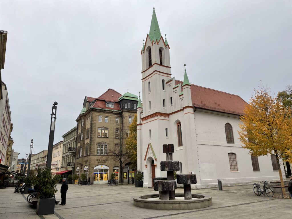 Die weiße Kirche mit grünem Turm steht am Altstadtplatz in Cottbus/Chóśebuz, umgeben von Herbstbäumen, historischen Fassaden und grauem Himmel.