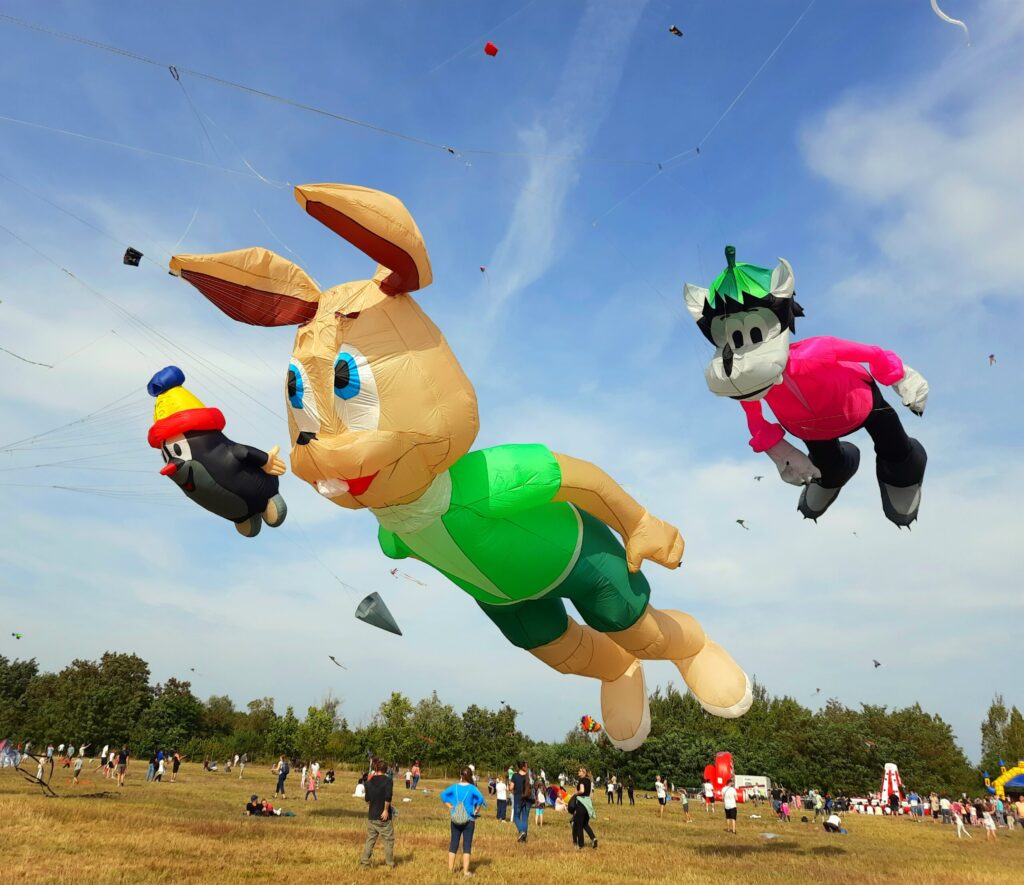 Mehrere riesige Drachen in Figurenform schweben über einer Wiese, während Besucher darunter bei blauem Himmel ein Drachenfest verfolgen.