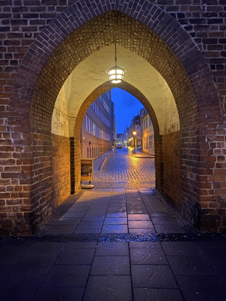 Gewölbter Backsteinbogen in einer Altstadtgasse bei Abendlicht, mit nassen Pflastersteinen, Laternen und historischen Fassaden im Hintergrund.