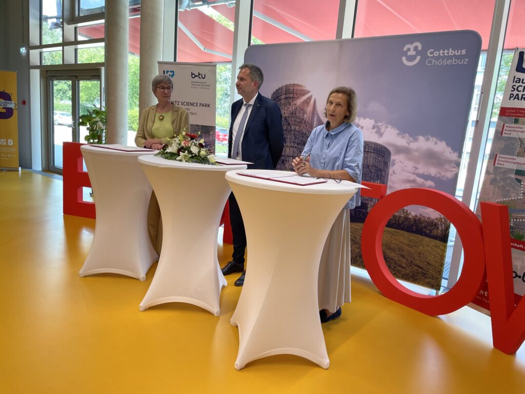 Im Cottbuser Science Park unterzeichnen Laura Kelly und weitere Vertreterinnen eine Vereinbarung an Stehtischen vor dem Stadtlogo im Innenraum.
